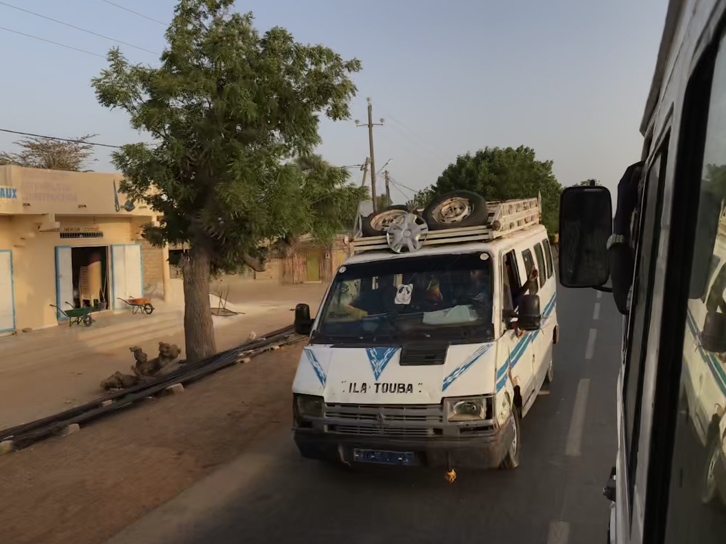 A white utility vehicle in Africa with blue stripes and writing, a damaged utility pole with fallen wires lying on the ground, a large tree, a building with a sign, and a wheelbarrow on the sidewalk.