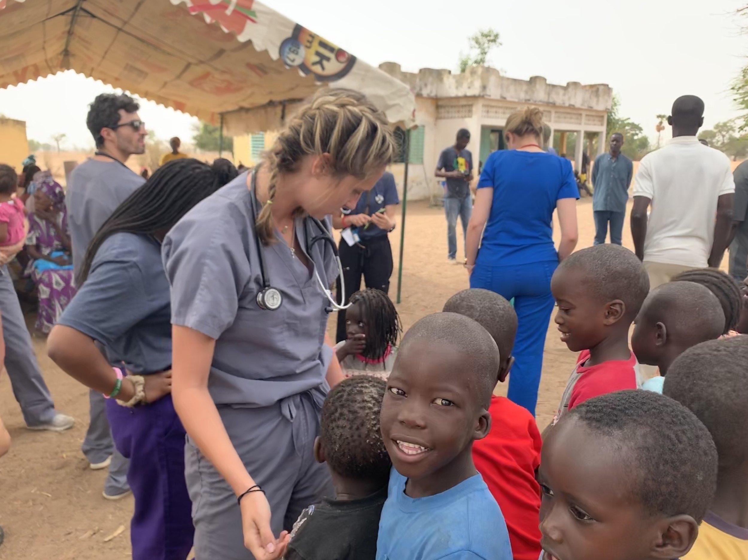 A group of medical volunteers, including a woman with a stethoscope, interact with children in an outdoor setting in a rural area, with some standing under a canopy and others on the ground.