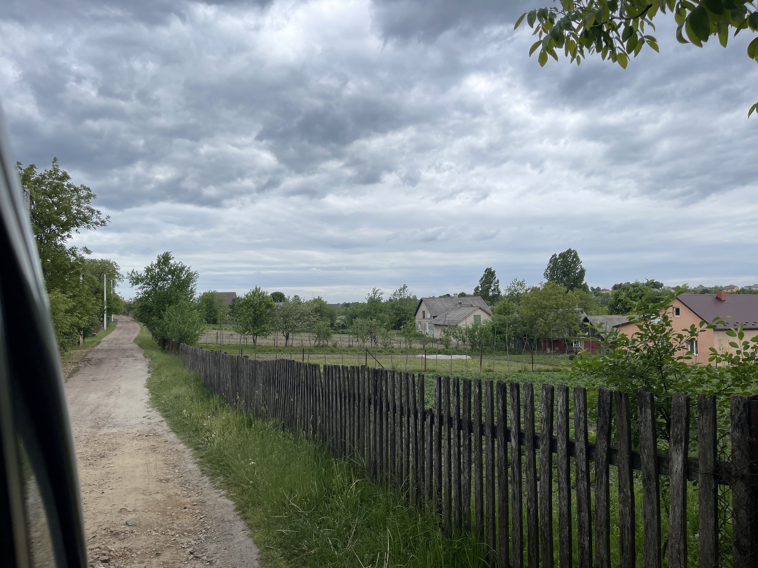A rural scene with a dirt road, green trees, a wooden fence, and houses in the distance under a cloudy sky in Ukraine during the Ukraine War