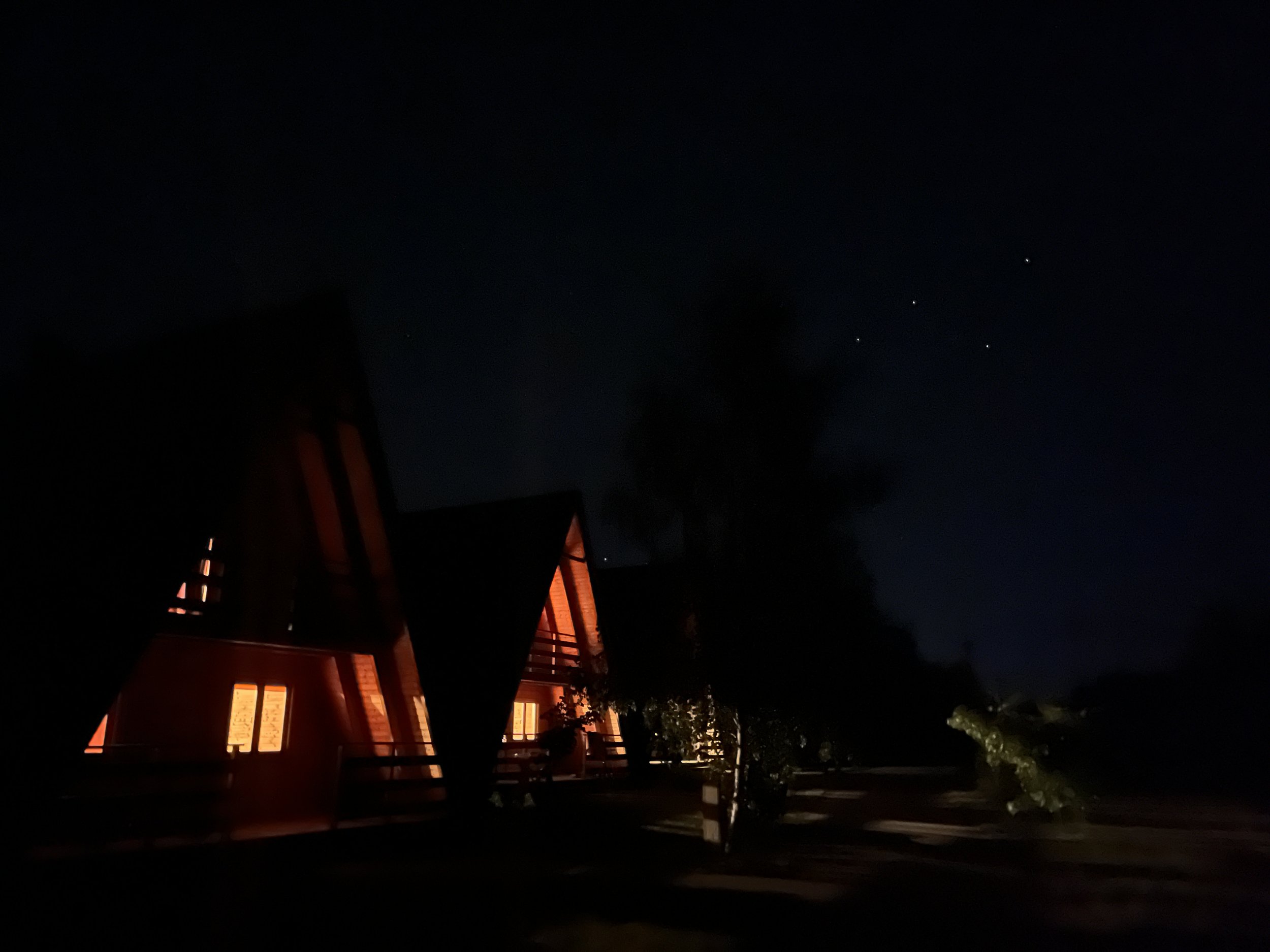 Night view of a building with illuminated windows and a balcony, with stars visible in the dark sky and trees in the foreground.