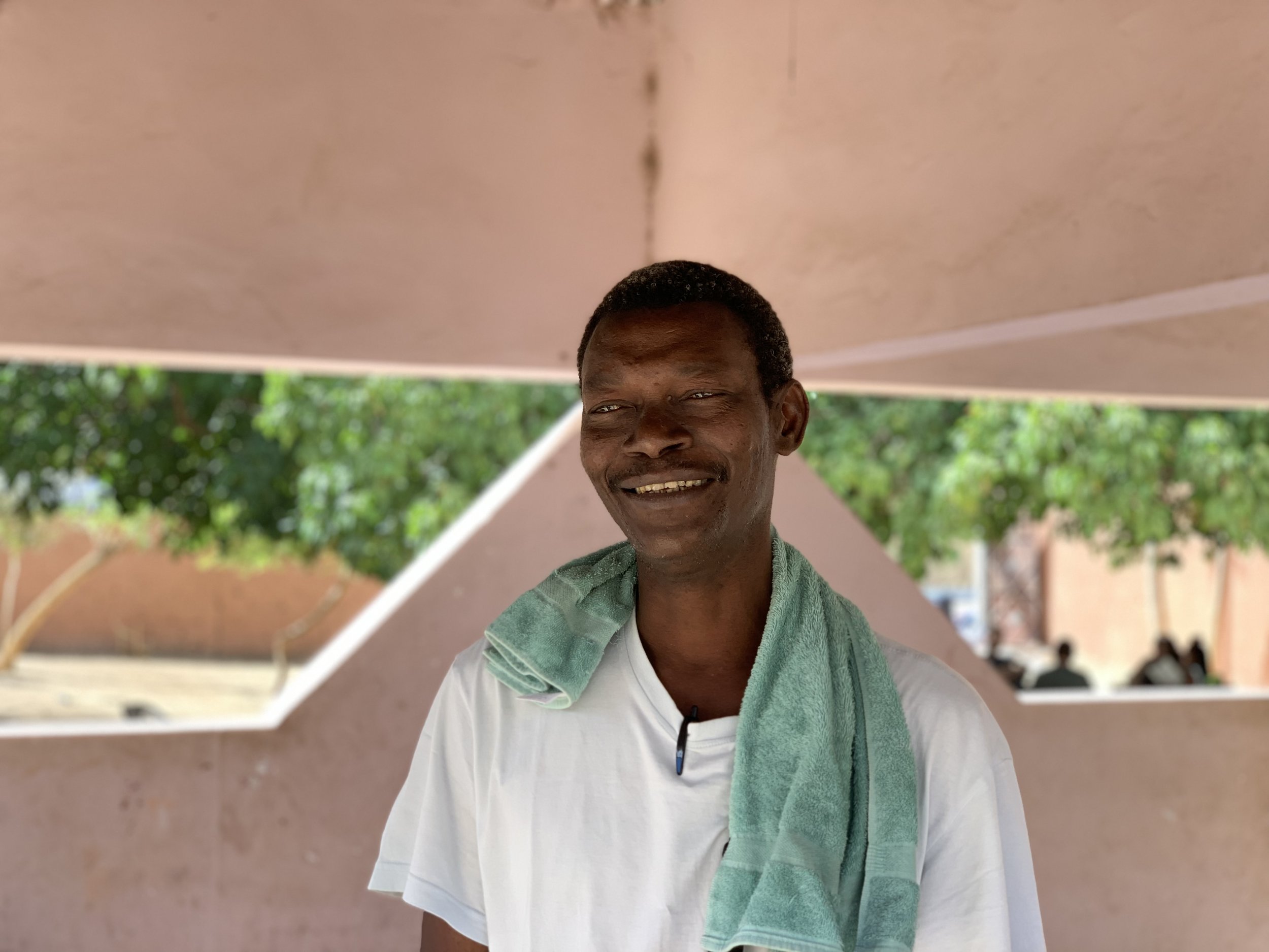 Smiling man with dark hair wearing a white t-shirt and a light green towel around his neck, outdoors with trees and a pink wall in the background.