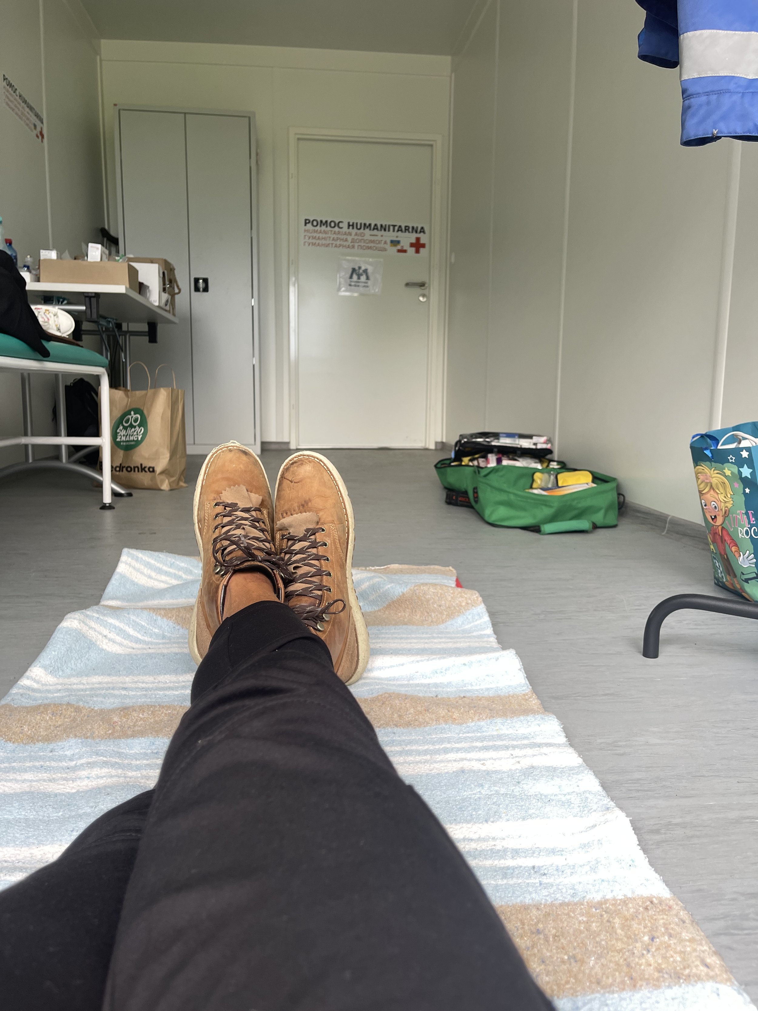 Person lying on a bed with legs stretched out, wearing black pants and brown boots, in a room with medical supplies, a green toolbox, and a door with a sign labeled 'Humanitarian Aid' in multiple languages.