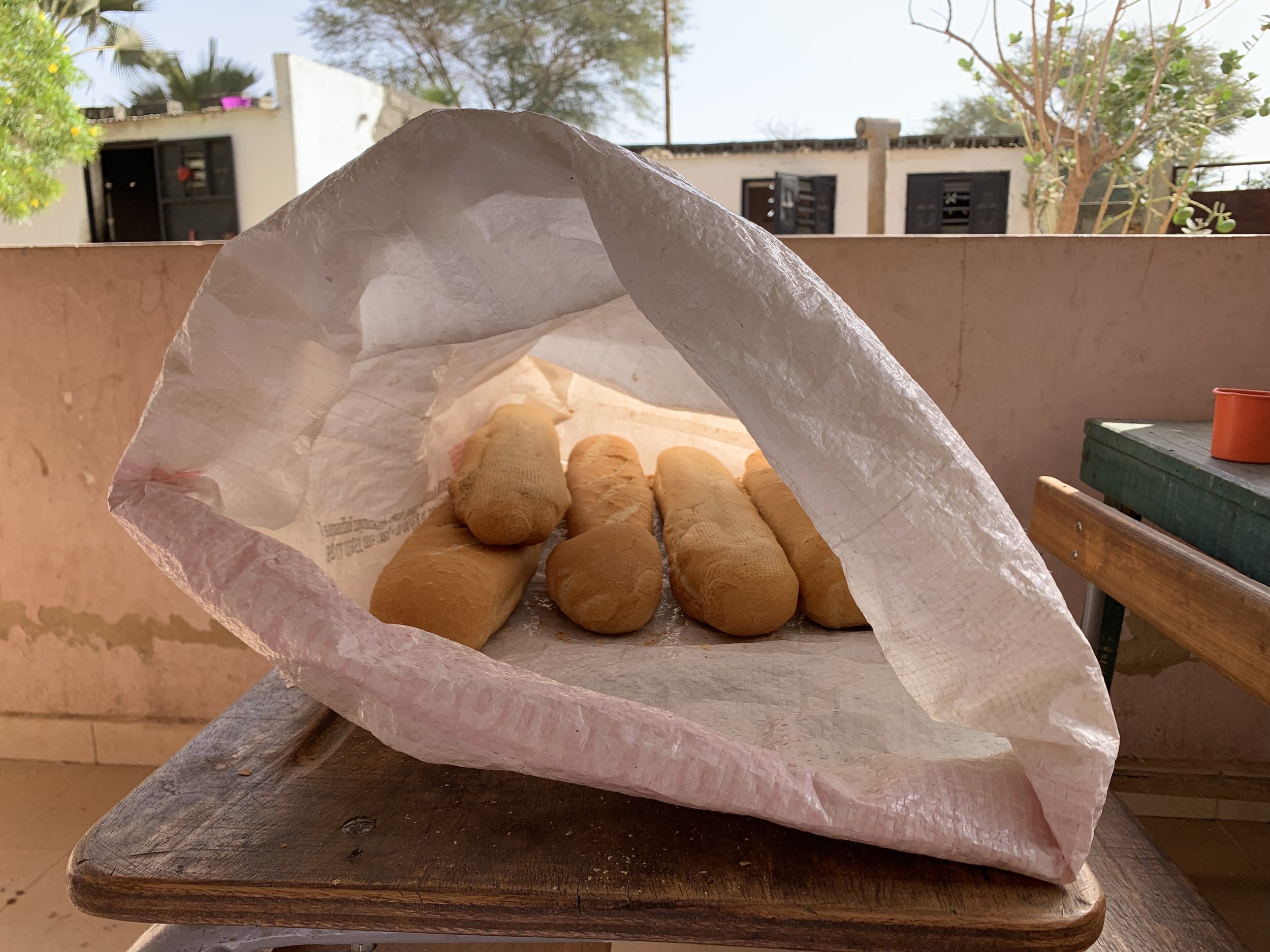 Baked bread rolls inside a paper bag on a wooden table, with an outdoor background including trees and buildings.