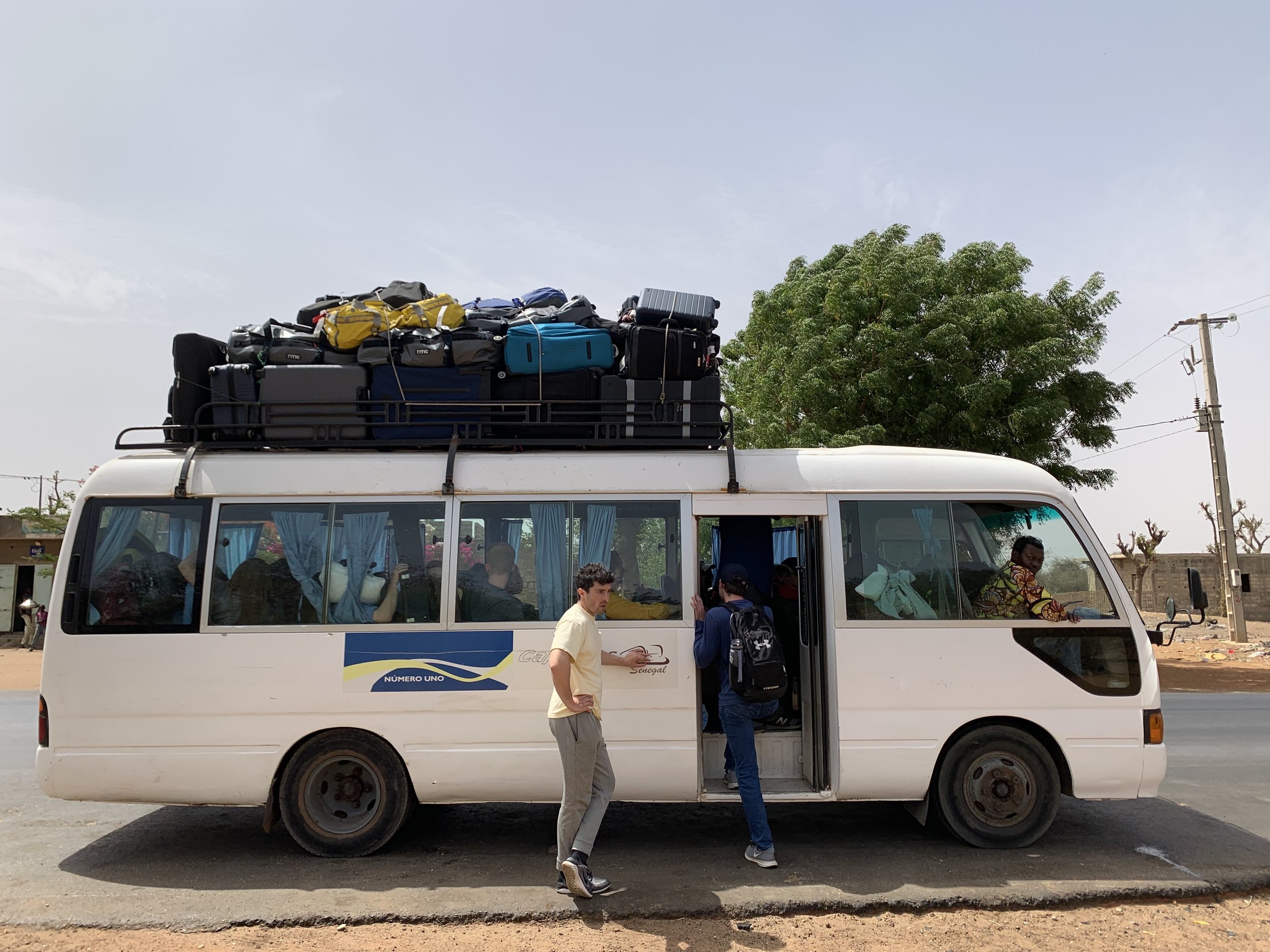 A white passenger bus in Africa parked on a street with luggage on the roof. Several people are near the bus, some inside and some standing outside. There is a tree and utility poles in the background.