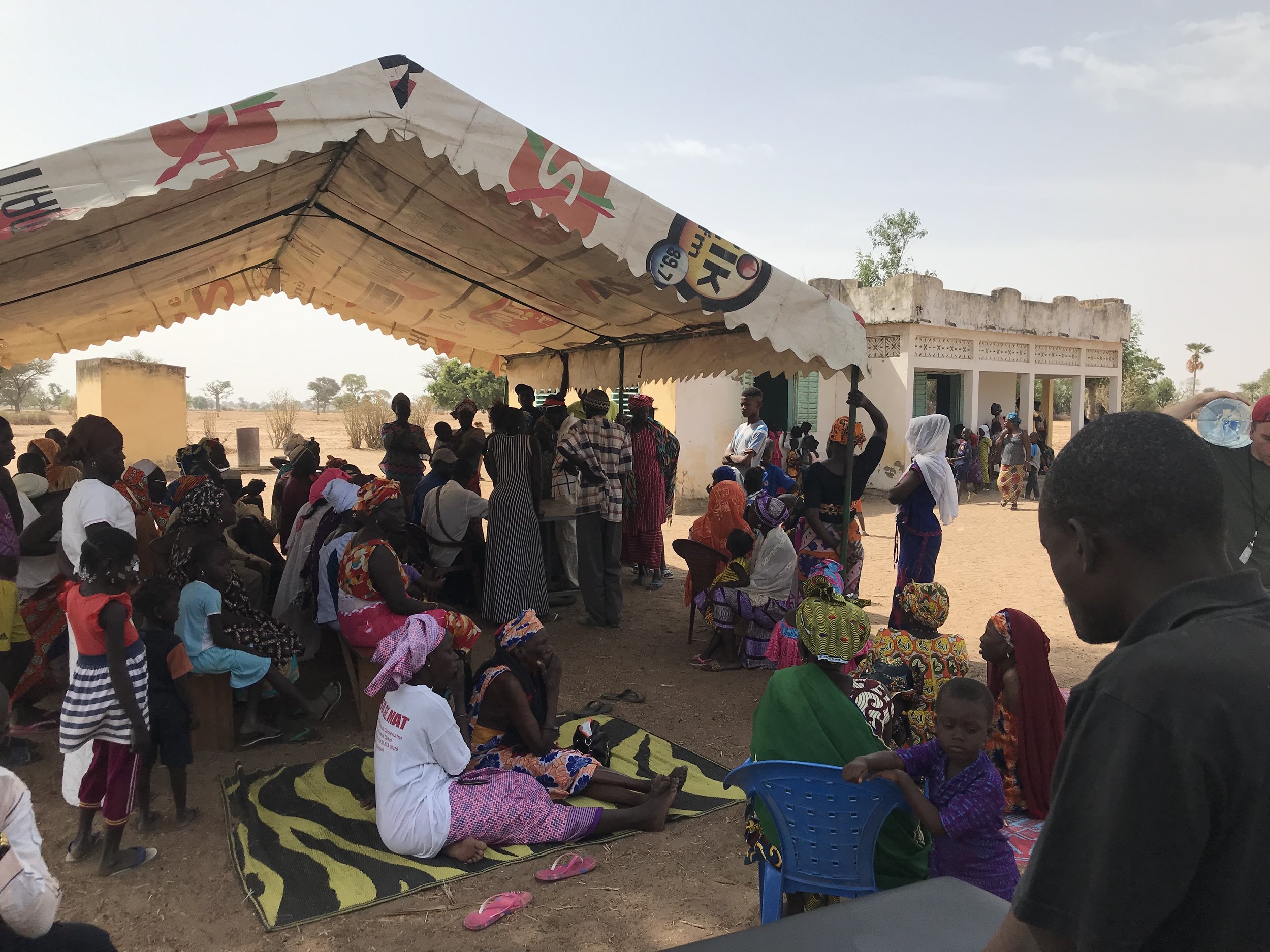 A large group of people gathered outdoors under a canopy in a rural area. The group includes men, women, and children, some sitting and some standing, dressed in colorful traditional clothing. The background shows a simple building, trees, and a clea
