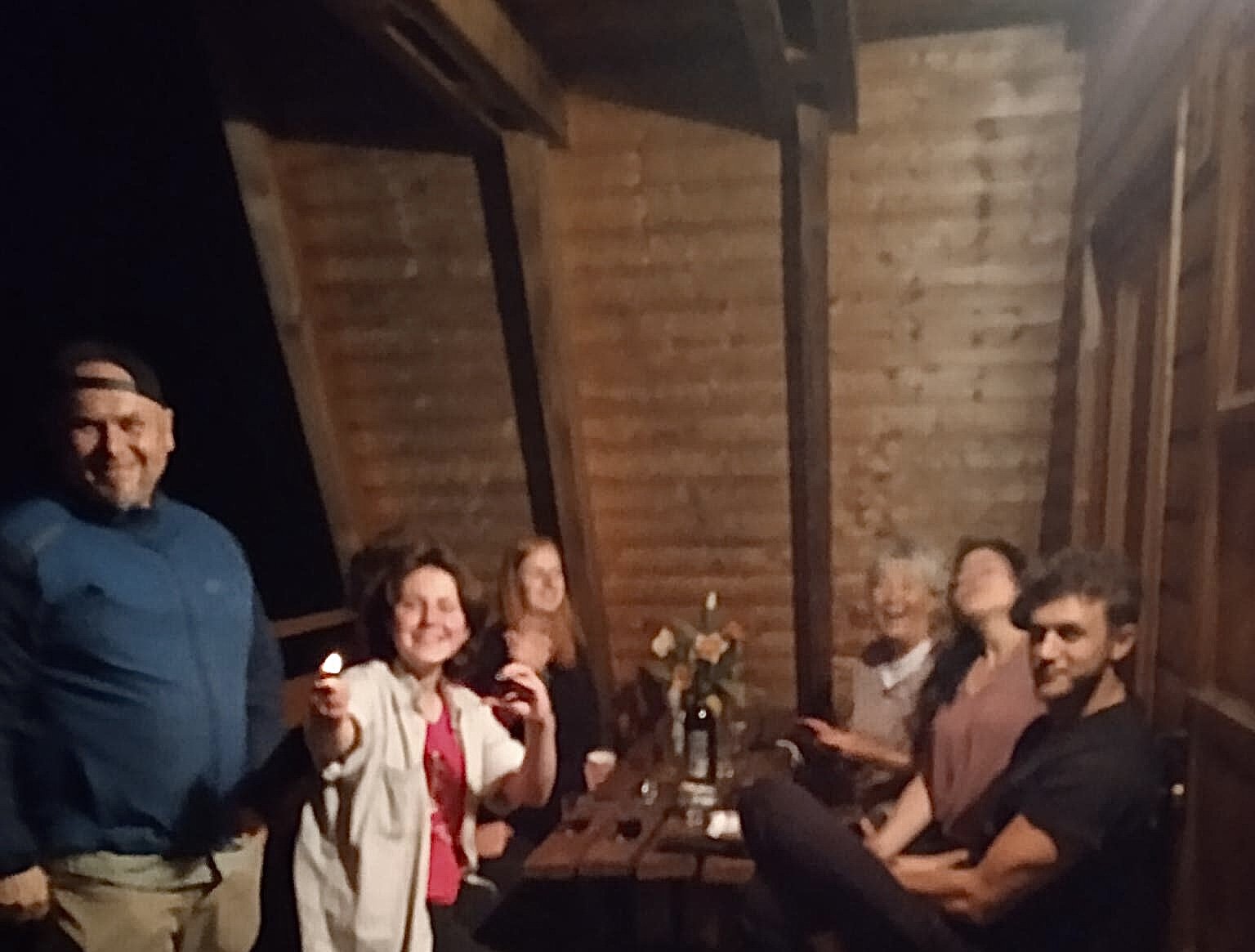 A group of five people sitting at a table in a wooden cabin, smiling and enjoying a social gathering in Ukraine during the war. There are candles and a flower vase on the table.