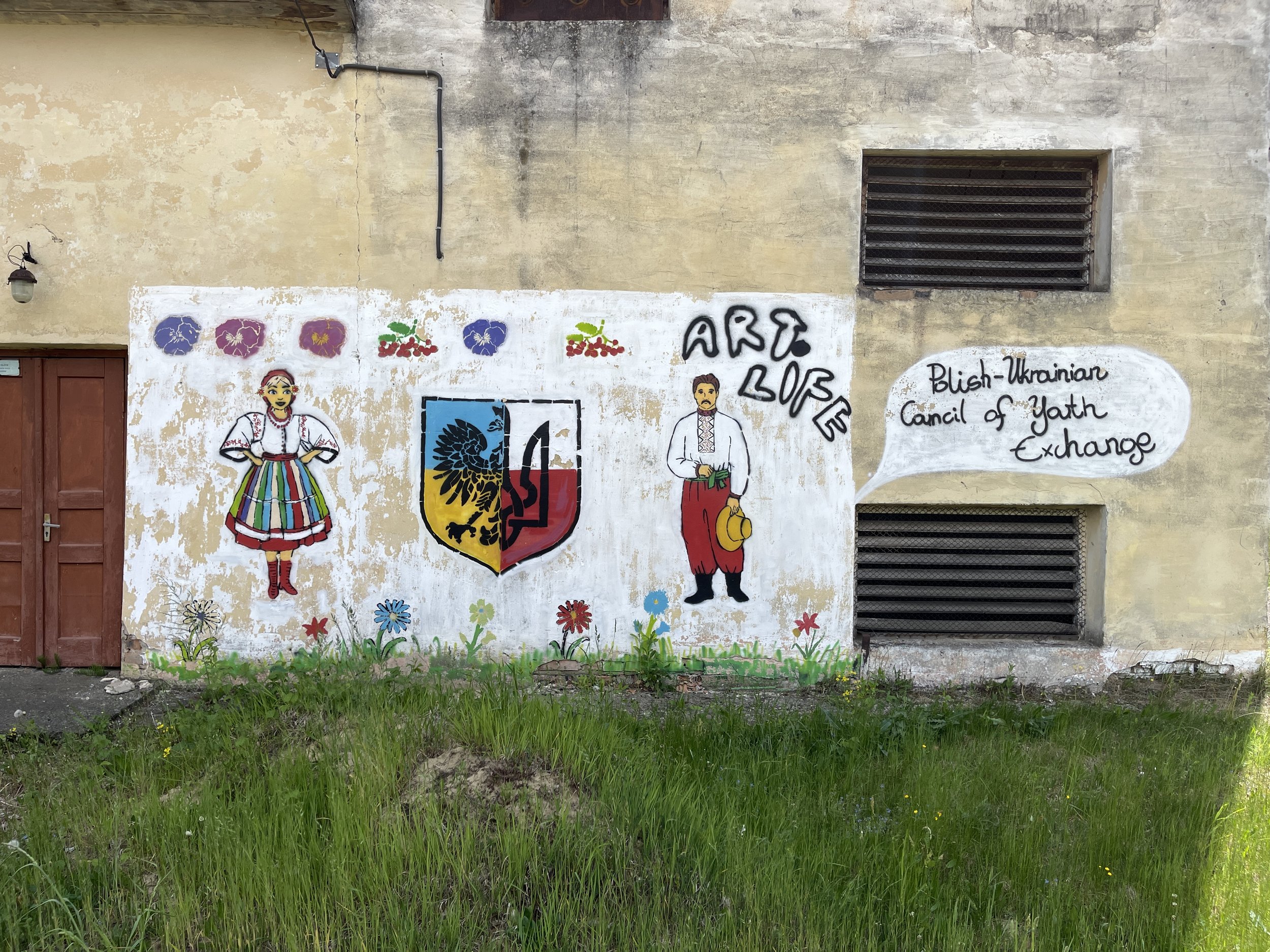 Muralon a weathered beige wall featuring traditional Ukrainian artwork, with two characters dressed in Ukrainian folk costumes, a woman in a floral dress and a man in embroidered attire, surrounded by colorful flowers and a shield emblem. A speech bubble states, "Blish-Ukrainian Council of Yath Exchange."