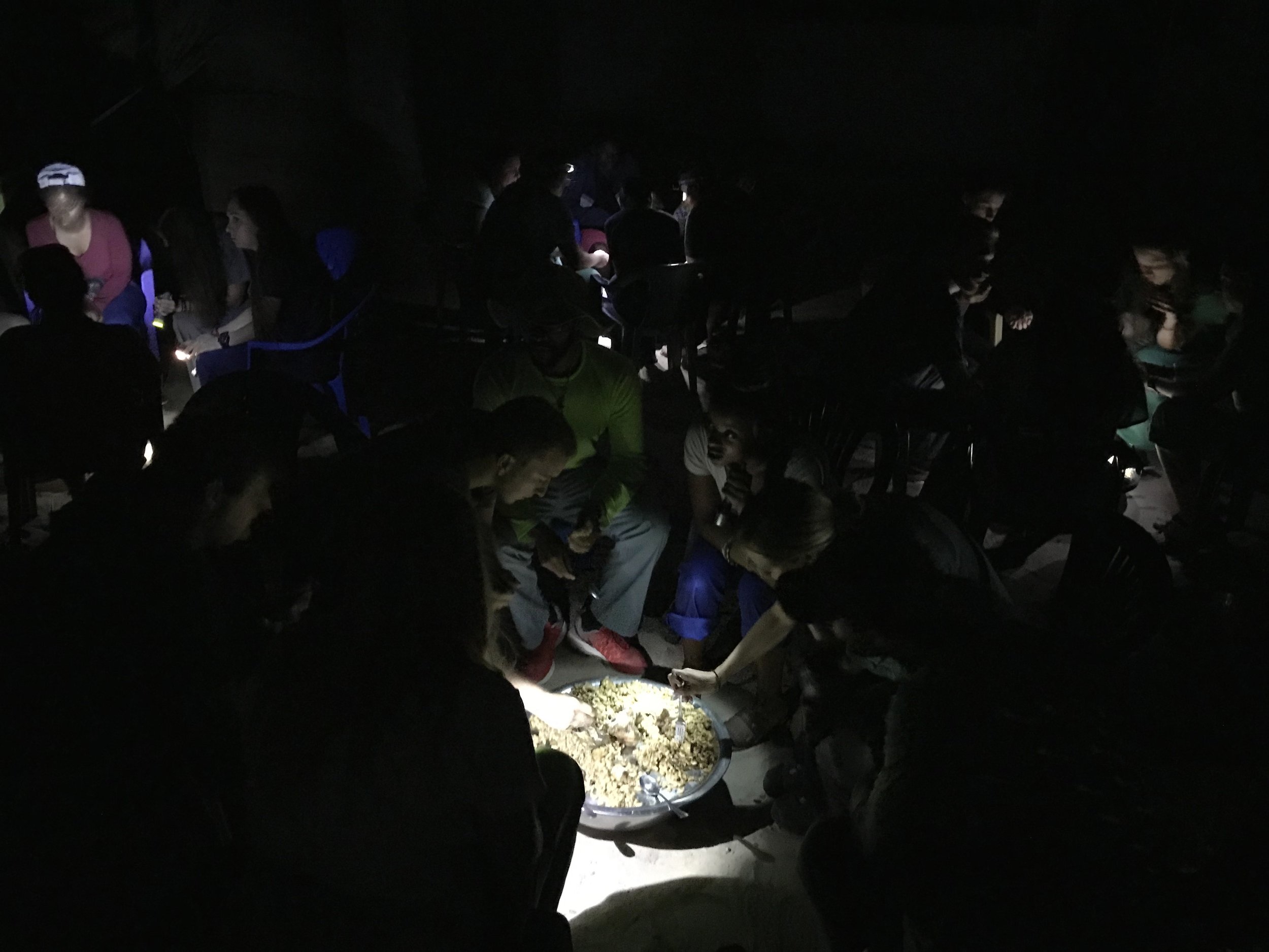 People gathered around a large dish of food under low lighting at night.
