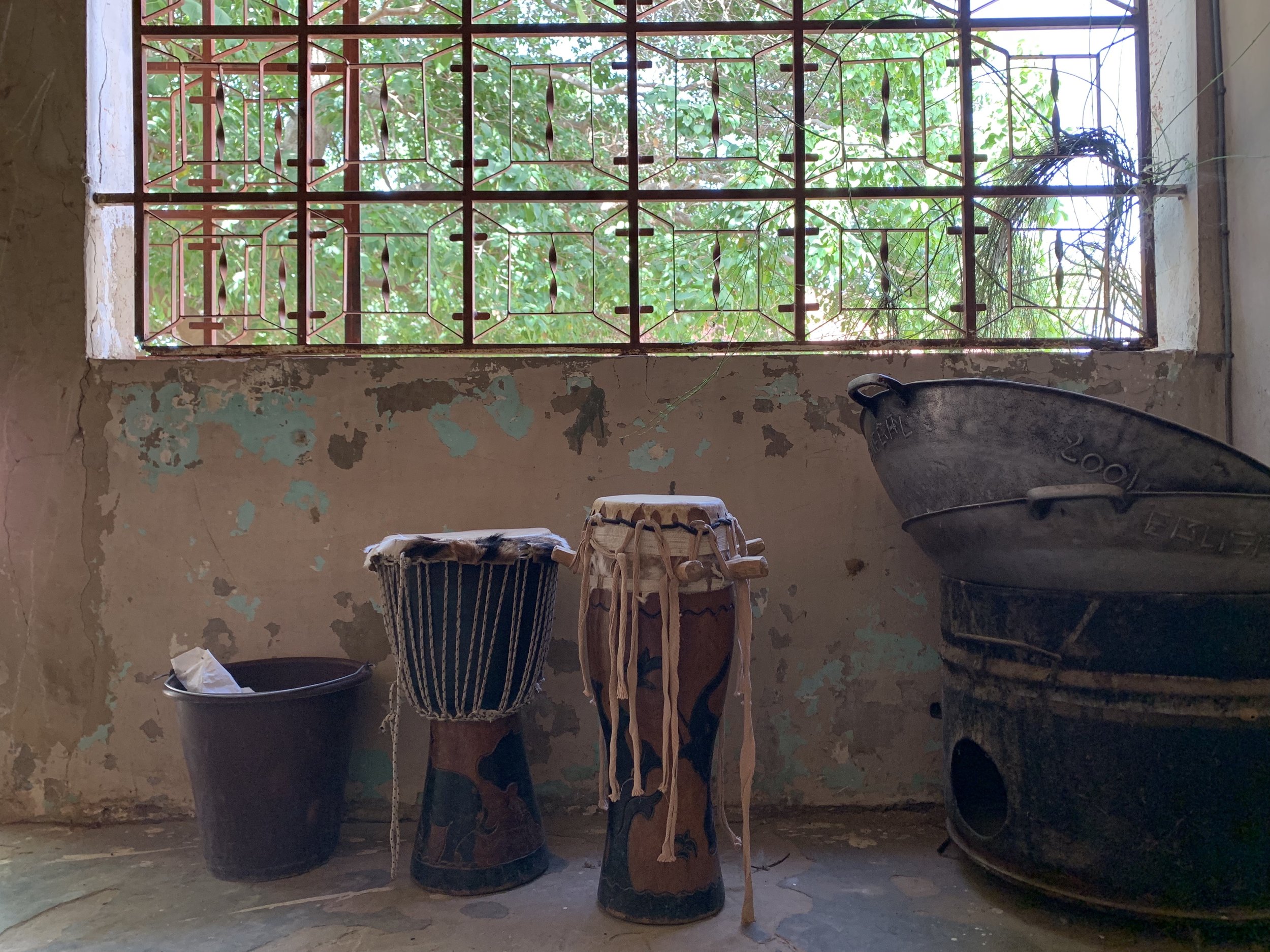 Interior of a rustic room in Africa with a large window featuring decorative iron bars, surrounded by peeling paint on the walls. There are various drums and buckets, some with cloth or paper covers, placed on the floor.