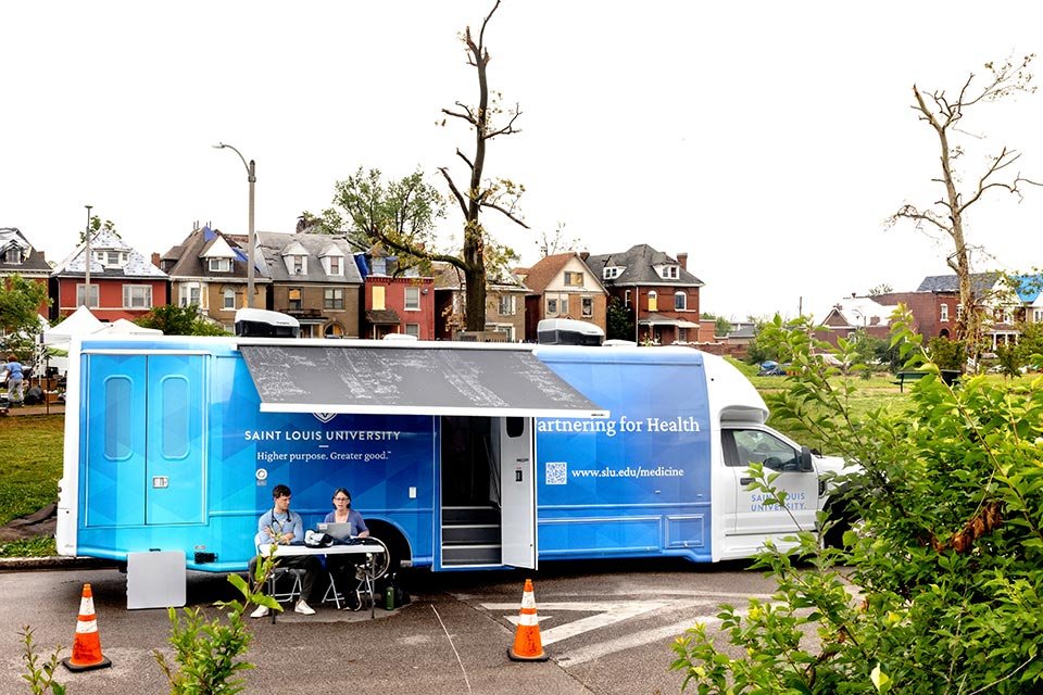 A mobile health clinic or outreach vehicle from Saint Louis University, parked outdoors with two people seated at a table, surrounded by some orange traffic cones and a lush green setting with trees and houses in the background.