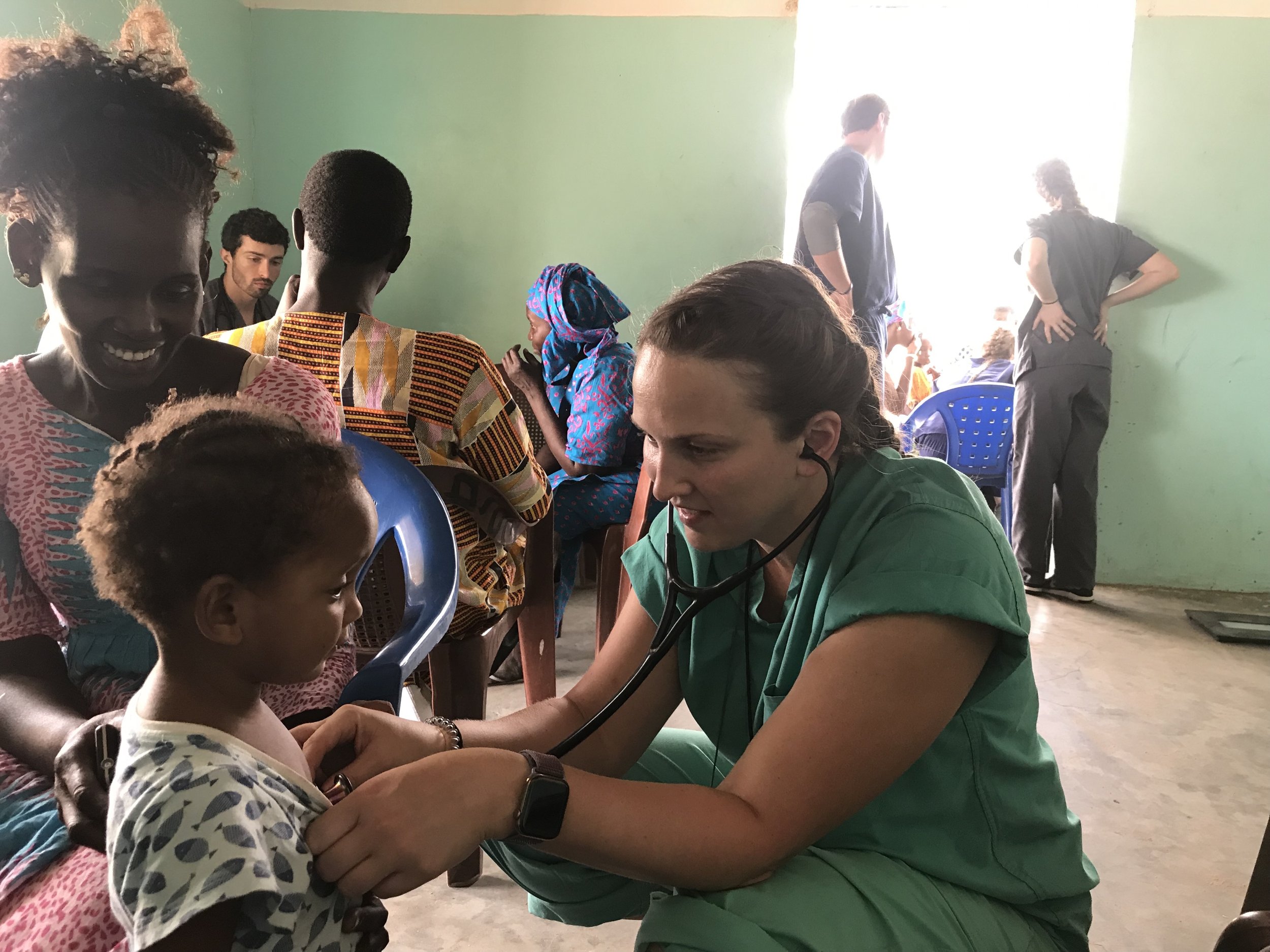 A healthcare worker in green scrubs using a stethoscope to check a young girl's heartbeat in a room with green walls and sunlight streaming in from a window. Several other people are present, including children and adults, some sitting and standing, 
