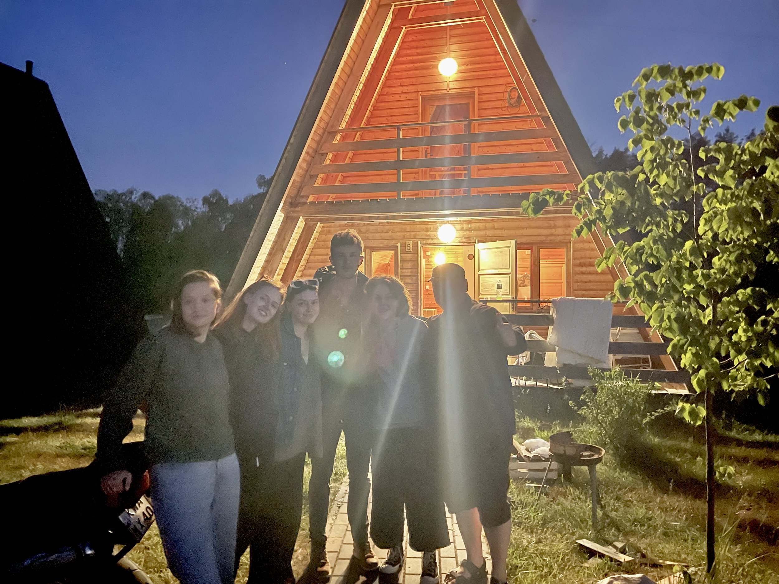 A group of people standing outdoors at night in front of a wooden A-frame house with lit windows and a porch, surrounded by greenery, in Ukraine during the war.