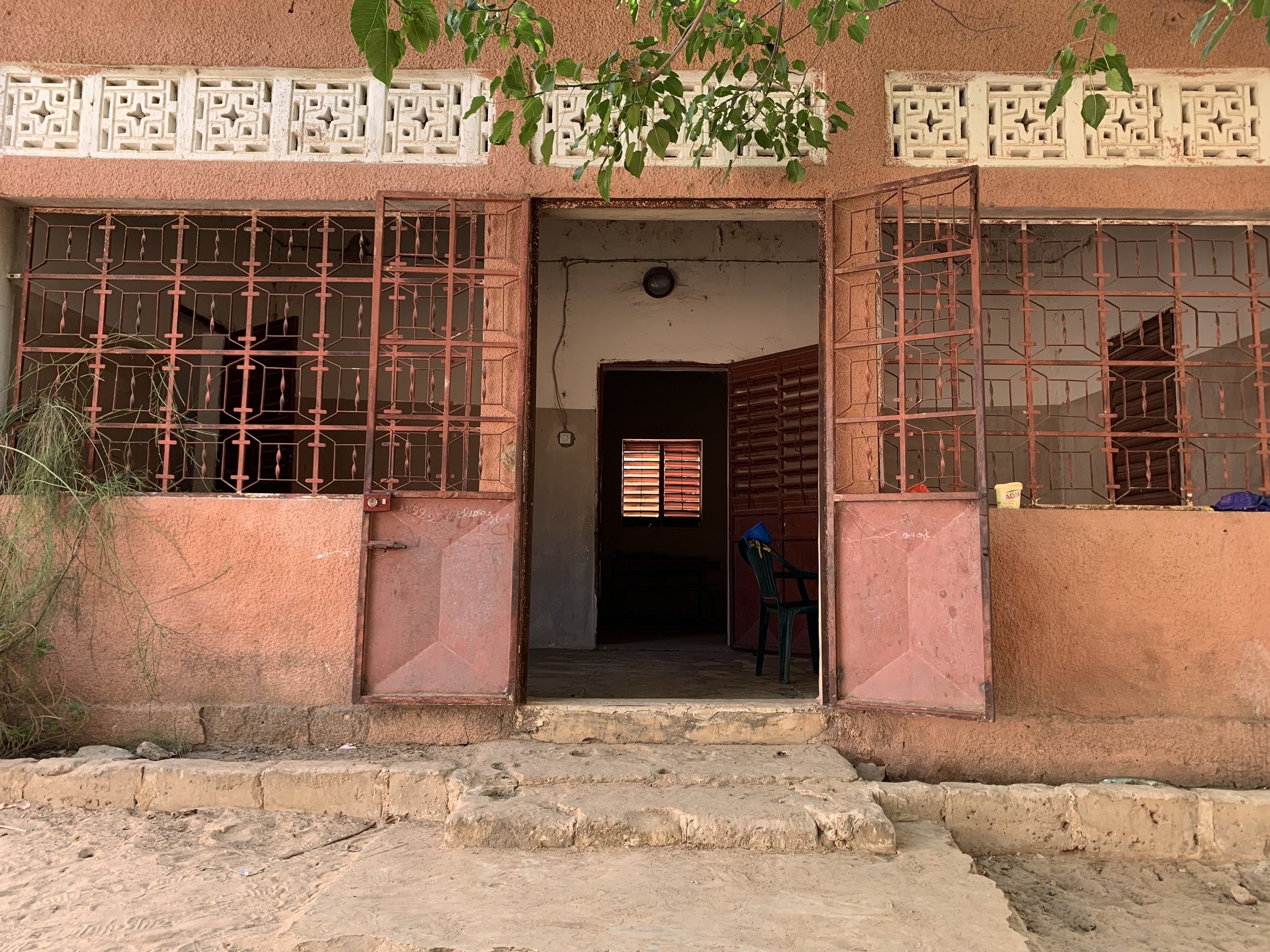 The image shows the front entrance of a building with an open door. The building has a pinkish wall with white decorative blocks near the roof. There are rusty metal gates on either side of the open door, and inside, there are stairs and a window wit