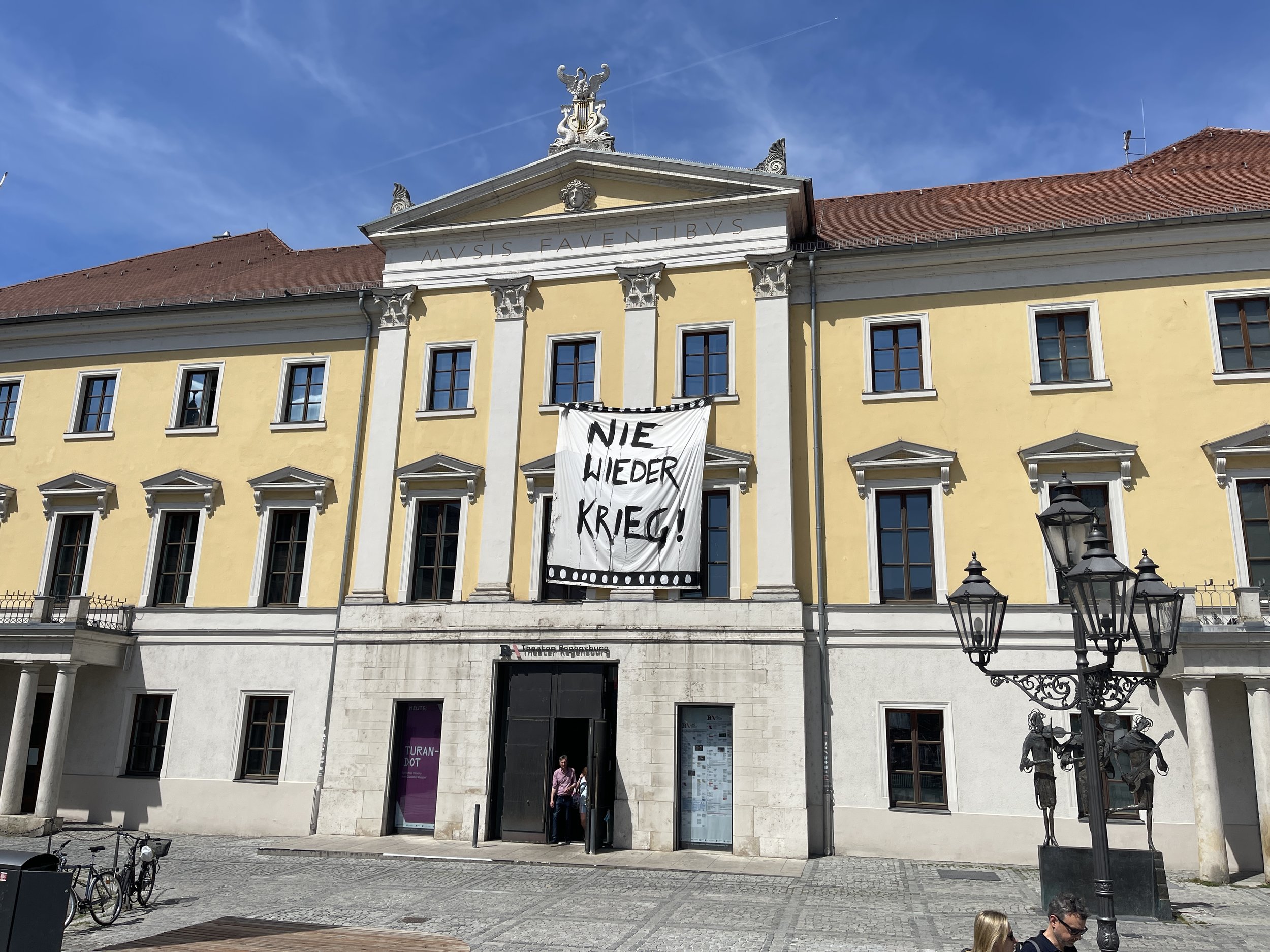 Historical yellow building in Regensburg Germany during the Ukraine War with white accents, a triangular pediment, and inscriptions, with people entering and bicycle parking in front; clear blue sky overhead.