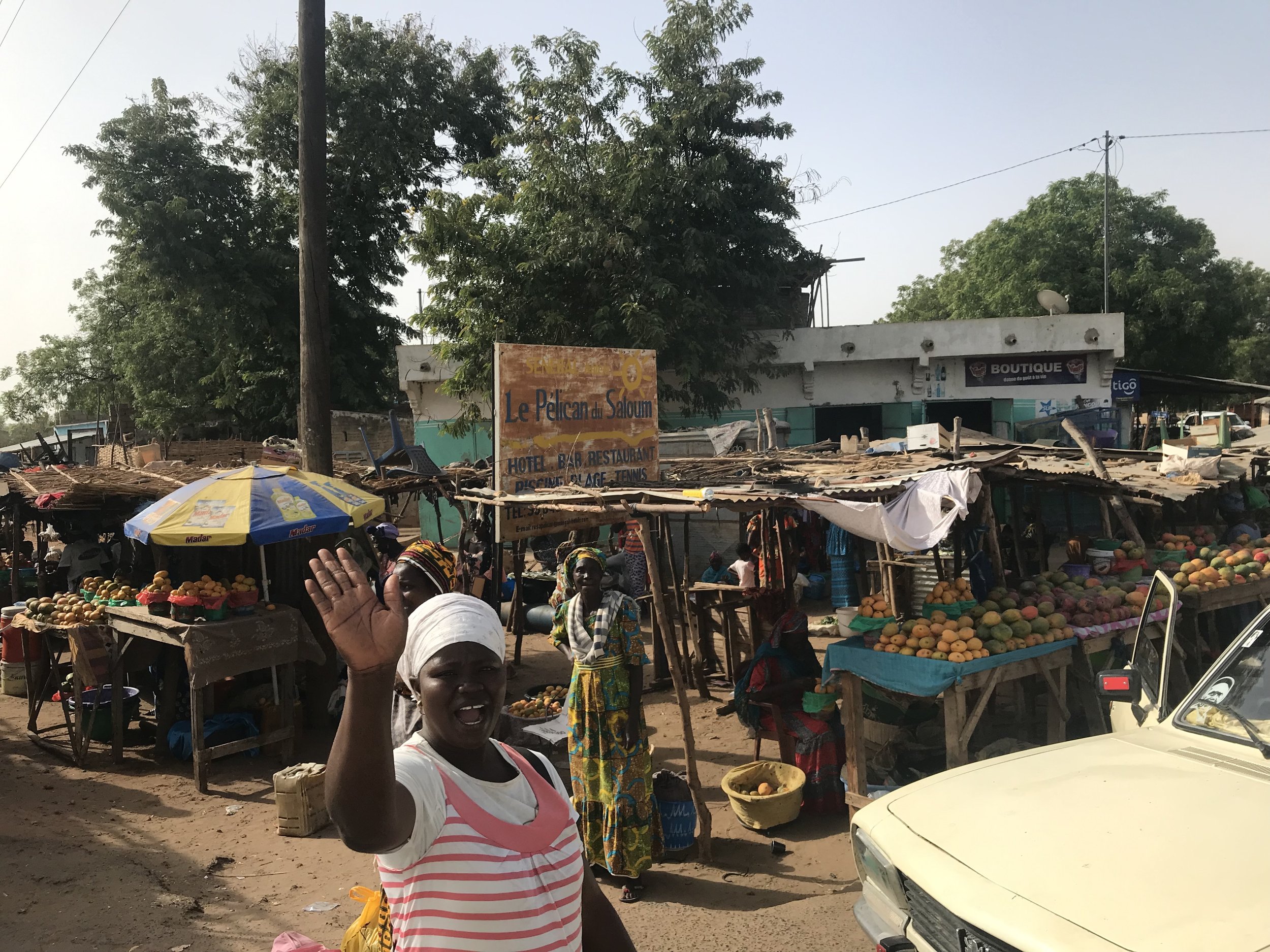 A woman waving at a bustling outdoor market, with vendor stalls selling fruits like oranges and tropical fruits, and signs indicating local businesses in the background.