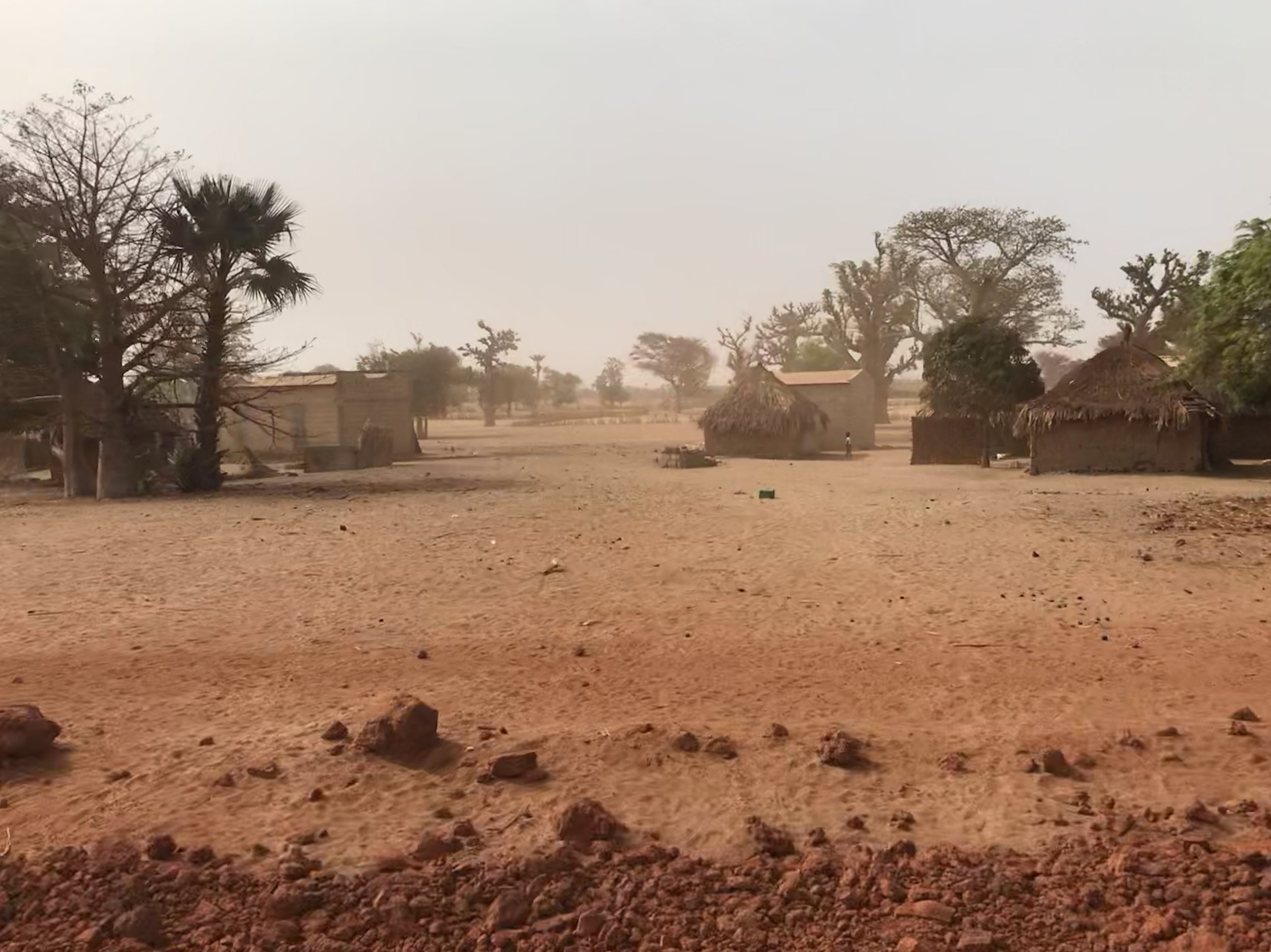 A rural village scene with traditional huts, trees, and dry, cracked ground in a dusty environment.