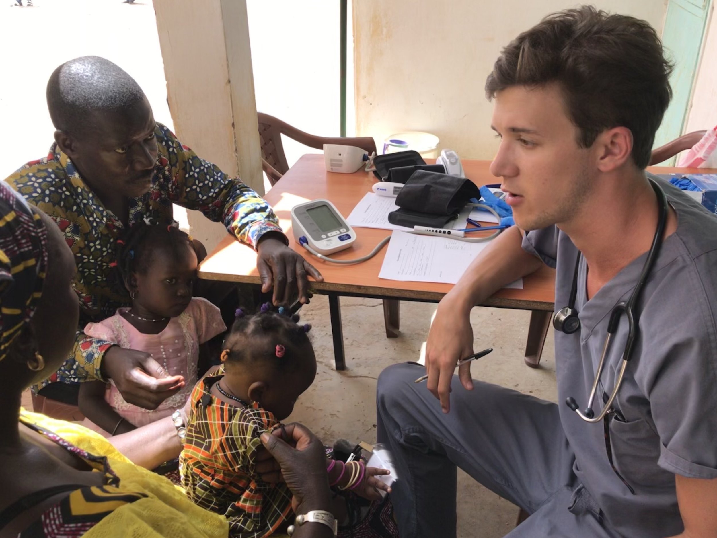 A healthcare professional, wearing a stethoscope and gray scrubs, sits at a desk talking to children and an adult woman in a rural setting. The children are sitting on the ground, and an adult woman with short hair, wearing a patterned dress, appears