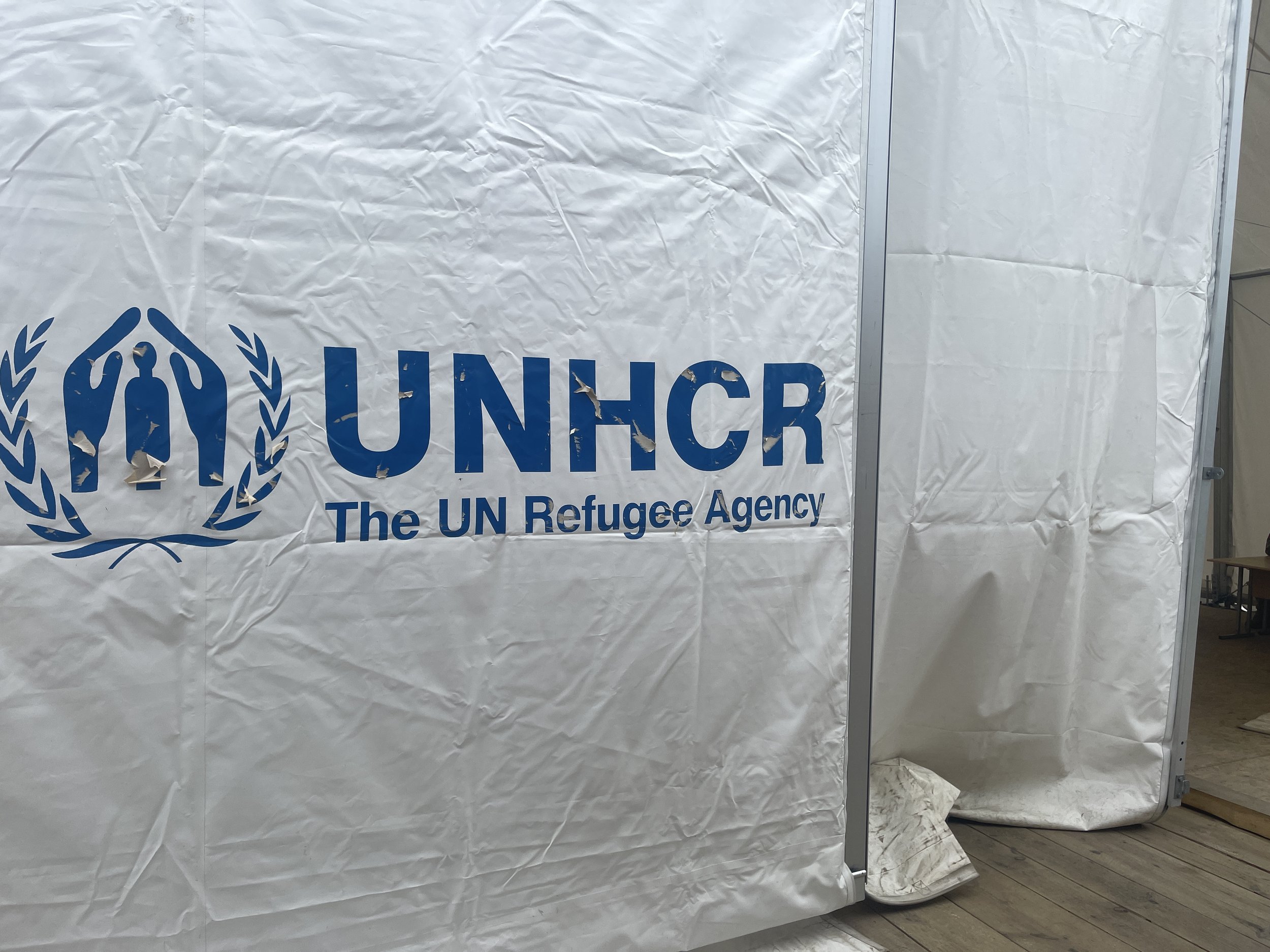 A white tent with the UNHCR logo and text, partially torn, with a metal pole in the foreground and tiled floor visible in the background.