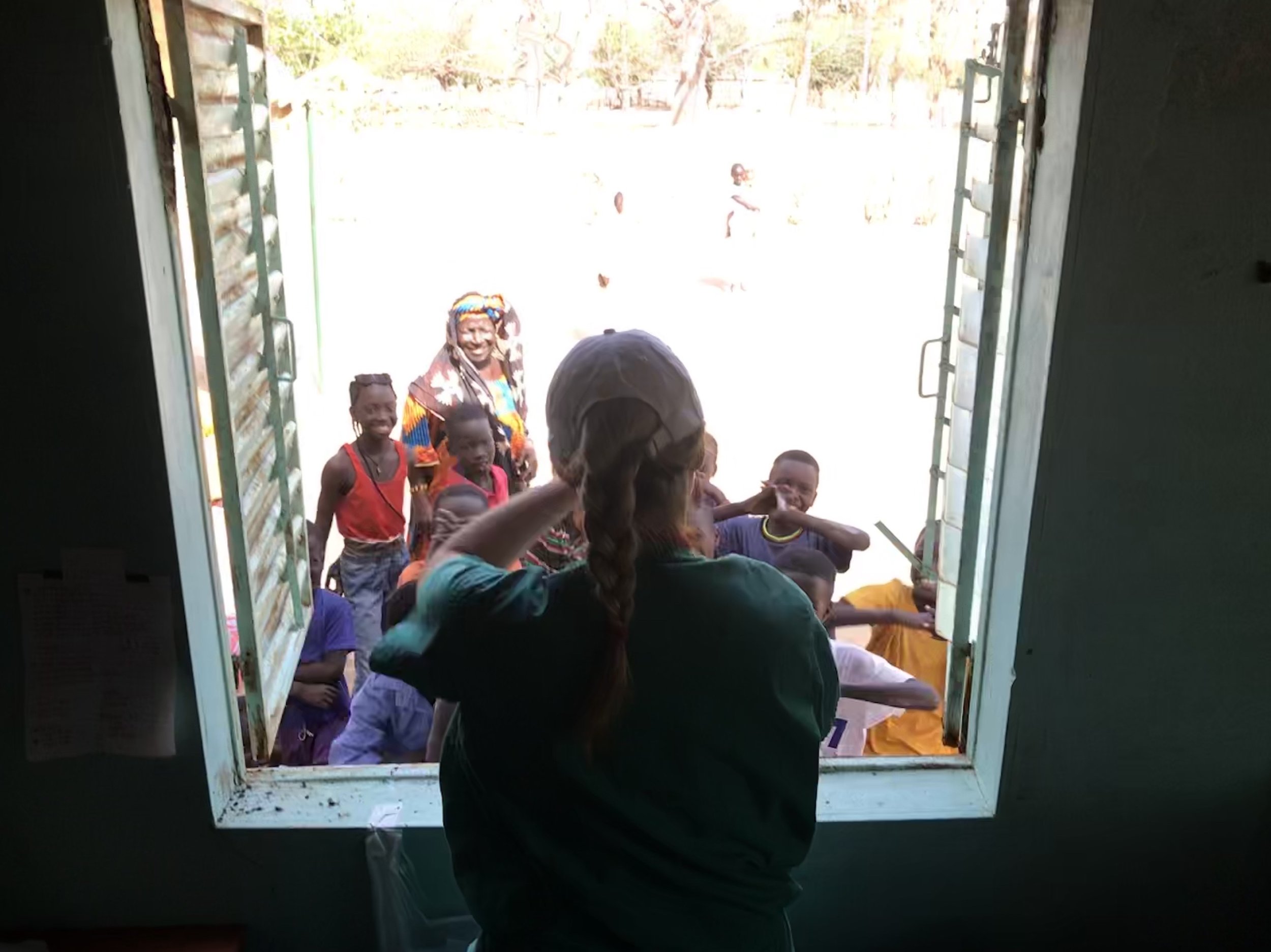 People, including children and an elderly woman, standing outside a building, seen through a window, with trees in the background.