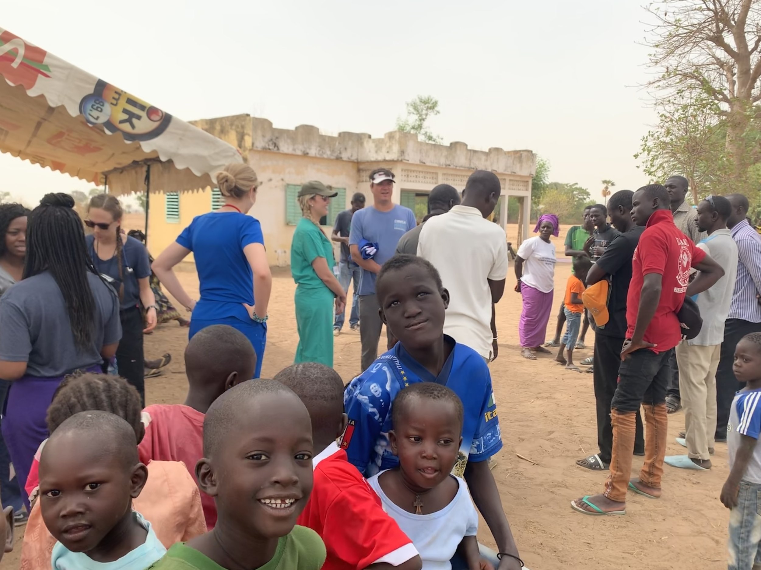 A group of children and adults gathered outdoors in a rural village setting, with some adults engaged in conversation and children looking at the camera.
