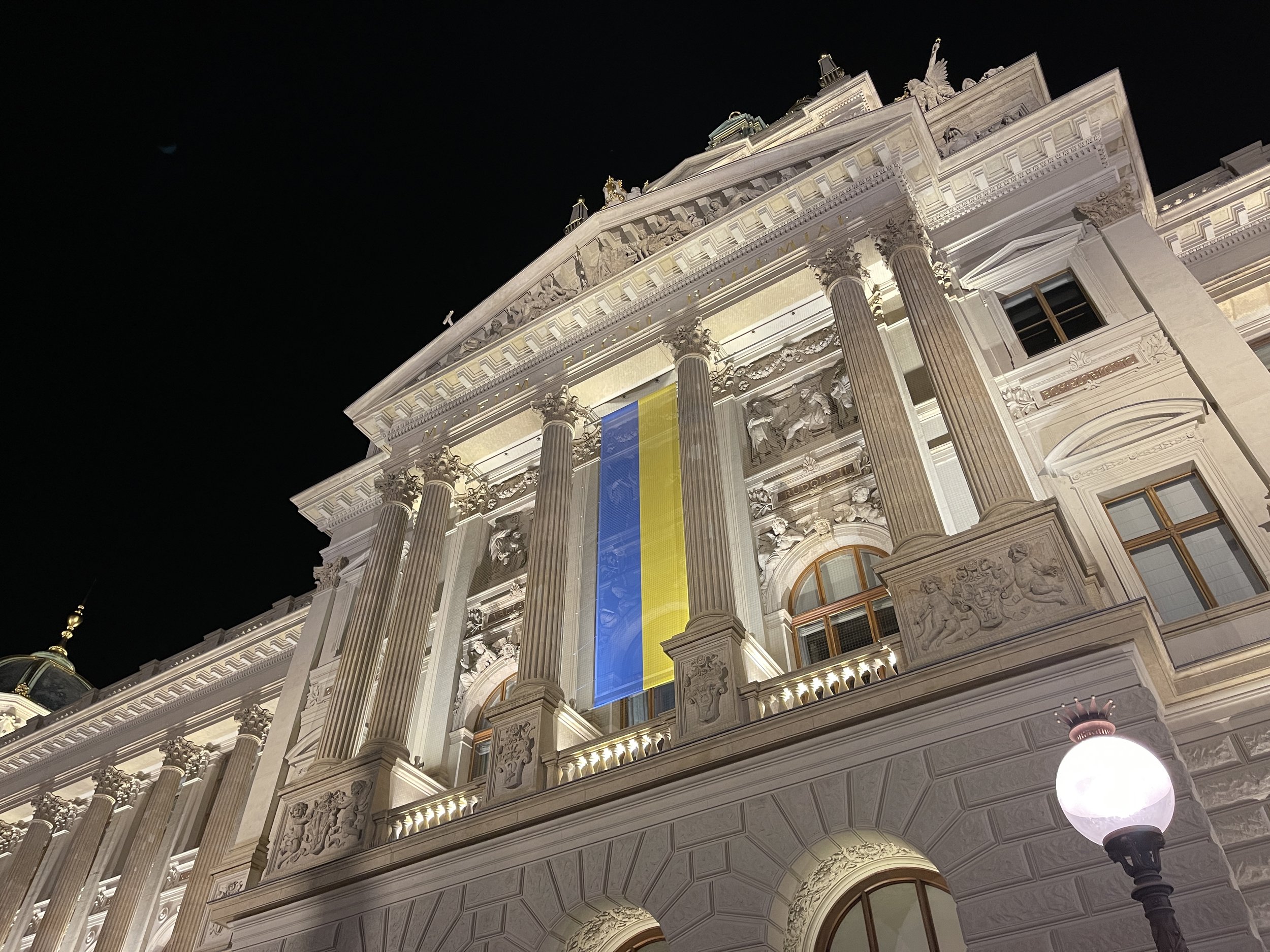 Night view of an ornate, classical-style building with tall columns, intricate sculptures, and gold accents illuminated against a dark sky, with a large blue and yellow banner hanging from the facade. Prague during Ukraine War.