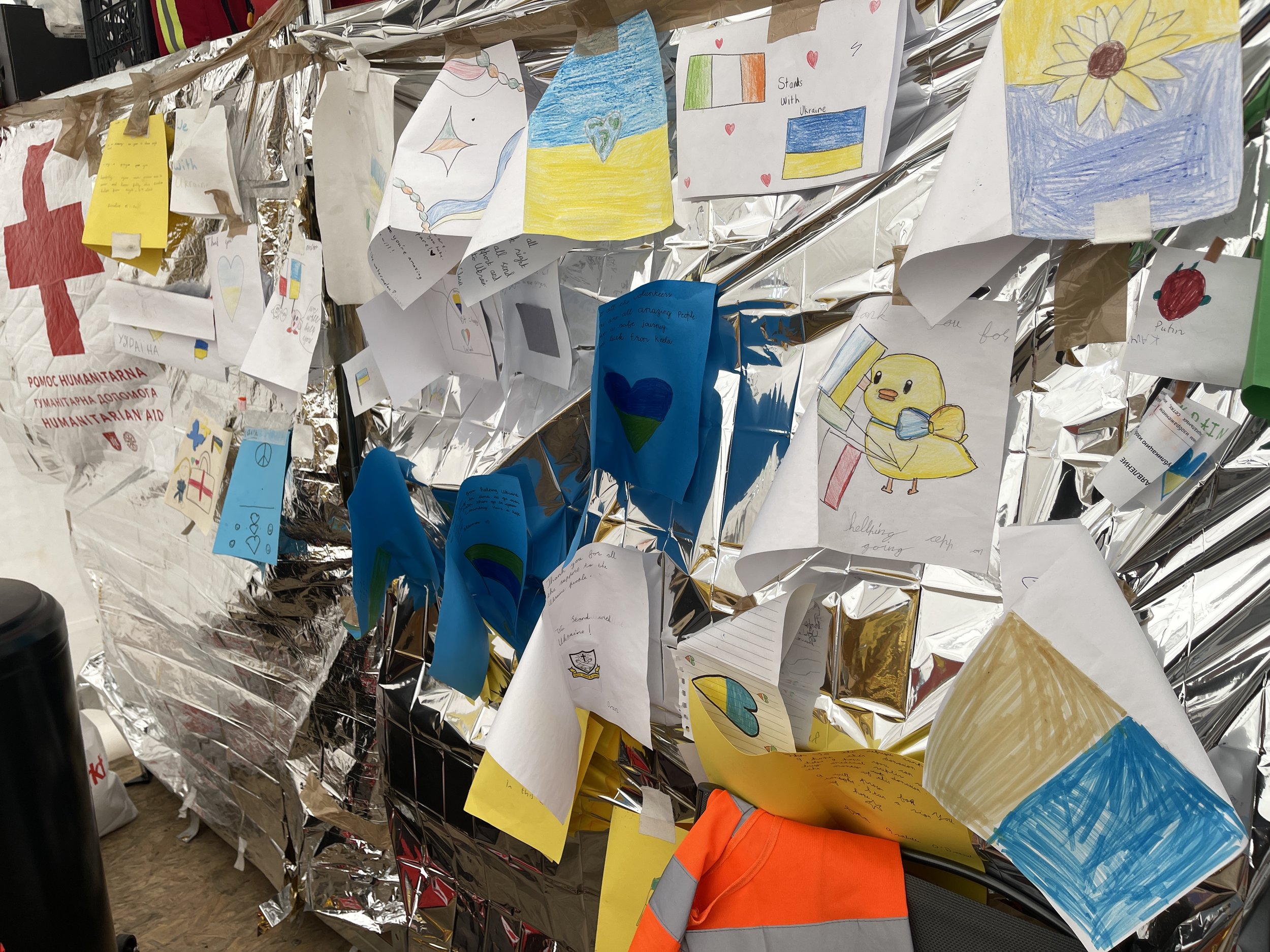 A collection of colorful drawings and handwritten notes pinned on a surface covered with crinkled foil, with some notes featuring flags of Ukraine. The drawings include representations of the Ukrainian flag, hearts, a chick, and flowers, with some no