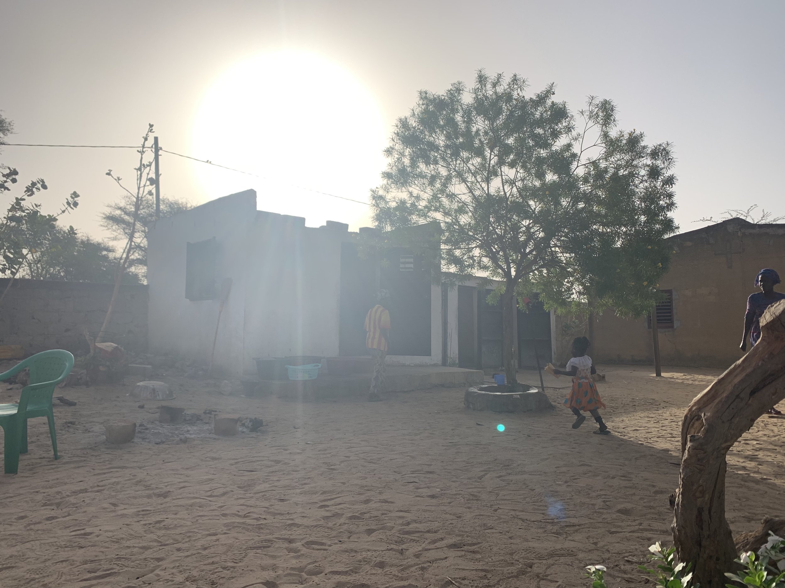A sunny day in a rural village with children playing outdoors, trees, a simple building, plastic chairs, and a dirt ground.