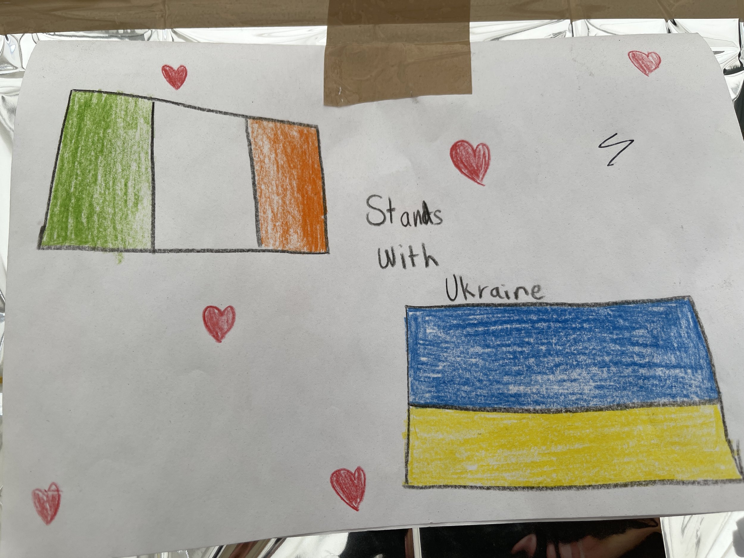 A hand-drawn picture of the Ukrainian flag with three horizontal stripes of blue, yellow, and blue, surrounded by red hearts.