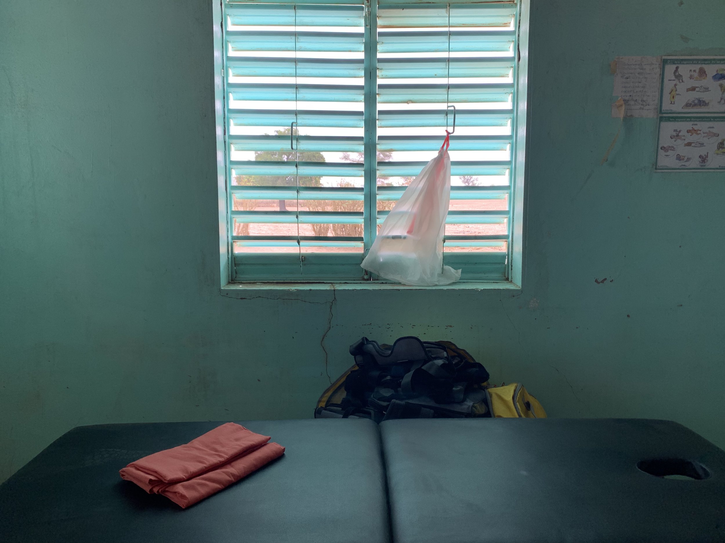 A small, simple room with green walls and a window with turquoise horizontally slatted blinds. There is a bed with a dark mattress and a folded pink blanket on top, positioned towards the bottom of the image. At the foot of the bed, there is a yellow