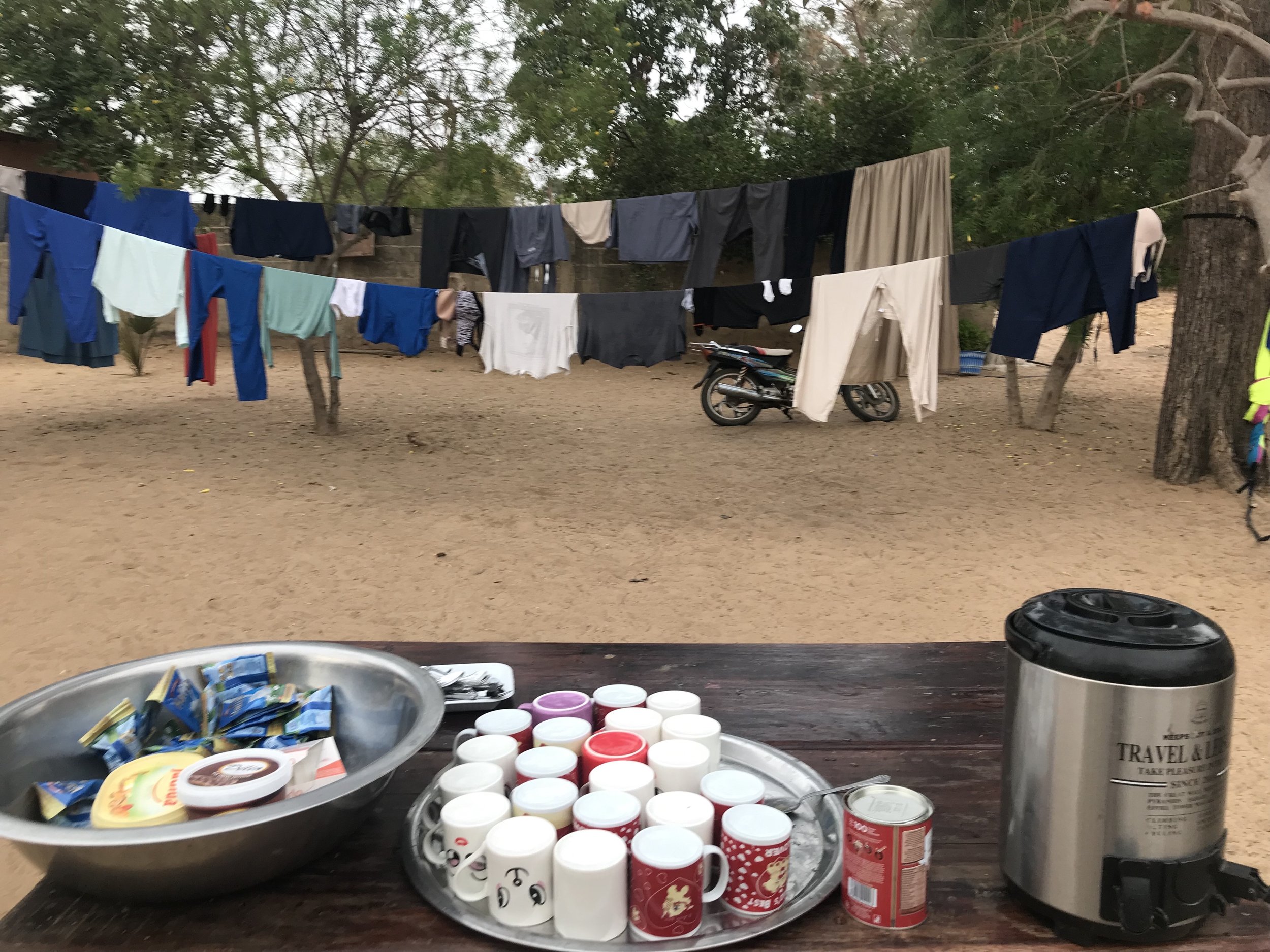 Table with various food items including cups, containers, and snack packaging, with clothes hanging on a line in the background and trees surrounding an outdoor area.