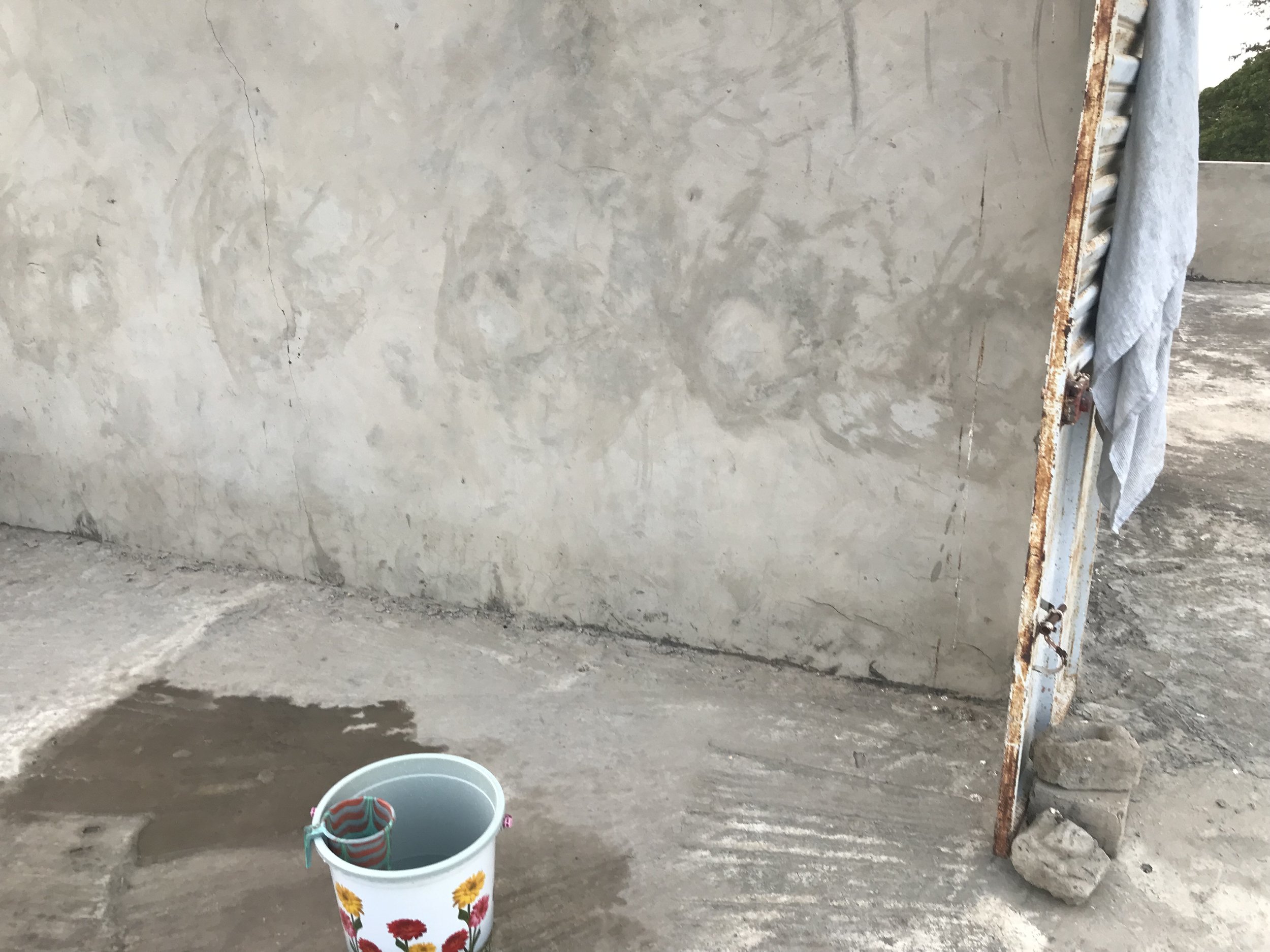 A concrete wall and floor in a construction site, with a rusted ladder leaning against the wall on the right side, and a white bucket with a colorful striped sock and a small tool inside on the ground in the foreground.